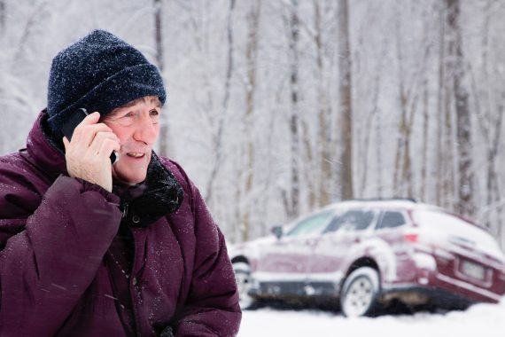 man on cellphone in snowstorm with car in snow bank on the side of the road
