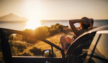 Person watching the sunset while reclining on the hood of their car with the door open
