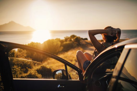 Person watching the sunset while reclining on the hood of their car with the door open